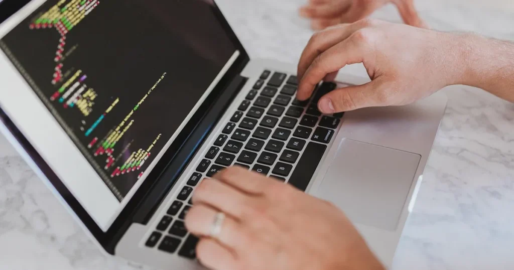 Deux mains tapent sur le clavier d'un ordinateur portable, du code affiché à l'écran.
