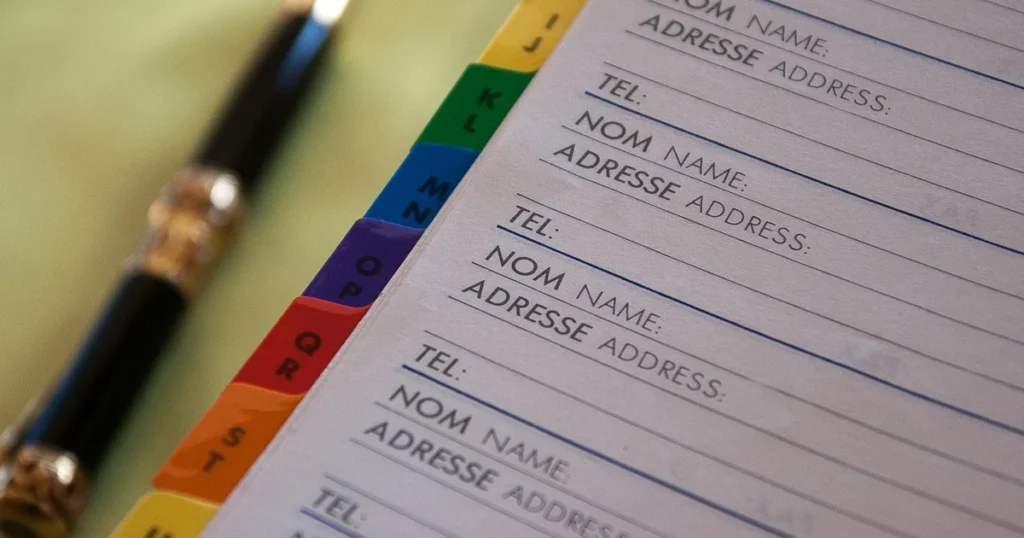 Un répertoire papier à onglets alphabétiques colorés, avec ses lignes Nom, Adresse et Tél., et un stylo posé à côté