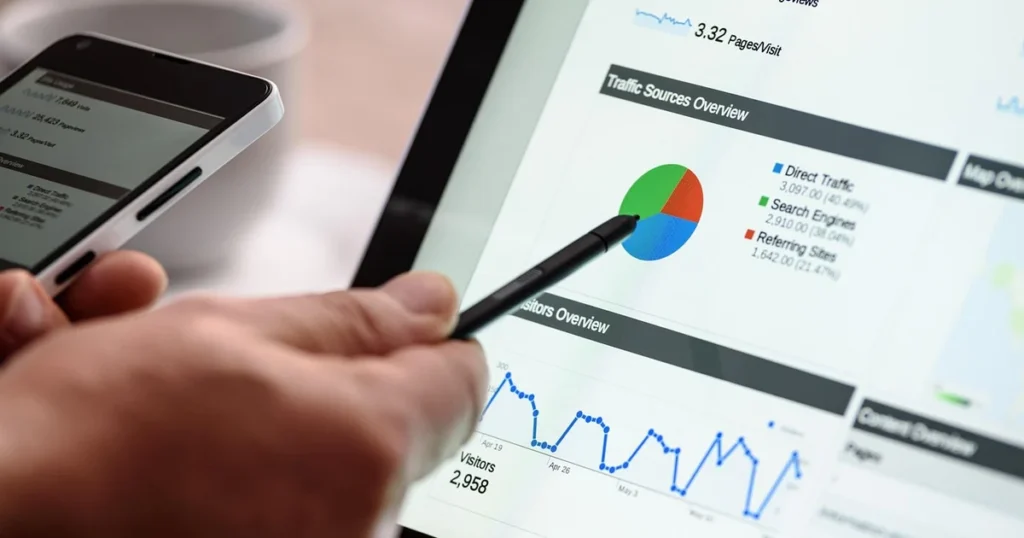 Une main tient un stylet devant un écran de statistiques de site : un graphique circulaire des sources de visites et une courbe de visiteurs, avec un téléphone à gauche